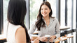 © NAMPIX - Smiling Asian businesswomen enjoying a coffee break and talking in a bright office cafe. Concept of positive office culture and friendship.