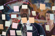 © TrueFrame Collective - Young adult Black man brainstorming ideas on transparent board covered with colorful sticky notes and diagrams, young adult Caucasian woman standing in background observing process