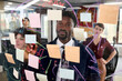 © TrueFrame Collective - Young Black man standing in front of transparent board with sticky notes, collaborating with young Caucasian women and drawing diagrams during creative brainstorming session in modern office