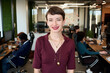 © TrueFrame Collective - Portrait of young adult Caucasian woman smiling confidently at camera in modern office workspace with diverse colleagues working at desks in background