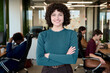 © TrueFrame Collective - Portrait of young adult Caucasian woman standing with arms crossed smiling at camera in modern office workspace with diverse colleagues working in background
