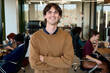 © TrueFrame Collective - Portrait of young adult Caucasian man smiling with arms crossed in modern office, diverse group of coworkers collaborating in background, open workspace environment visible