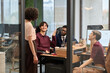 © TrueFrame Collective - Diverse group of young adult men and women collaborating in modern office, Caucasian man gesturing while talking to standing Caucasian woman, Black man and Caucasian woman listening