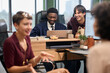 © TrueFrame Collective - Group of young adult multiethnic colleagues collaborating around desk, Black man and Caucasian man smiling while working on laptop, two women gesturing and talking in foreground