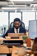 © TrueFrame Collective - Young adult Black man sitting at desk working on laptop in modern office environment, focusing on computer screen with charts visible on monitor, surrounded by office supplies