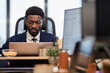 © TrueFrame Collective - Young adult Black man sitting at desk working on laptop in modern office environment, focused on screen with digital devices and office supplies visible in foreground