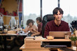 © TrueFrame Collective - Young adult Caucasian man working on laptop at modern office desk, focusing on screen with colleagues collaborating in background, stack of books and potted plant on workspace