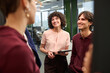 © TrueFrame Collective - Young adult Caucasian woman holding digital tablet standing with young adult Caucasian man smiling and listening to colleague during informal meeting in modern office workspace