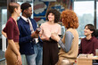 © TrueFrame Collective - Group of young adult multiethnic colleagues standing and talking in modern office, Black man holding coffee cup, Caucasian woman holding mug, everyone smiling and interacting