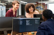 © TrueFrame Collective - Two young adult Caucasian women smiling and leaning over computer monitors engaging in conversation with young adult Black man sitting at workstation in modern office setting
