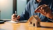 © alvi - Close-up of a man's hands holding a small house model on a wooden desk with notepad and pen in a blurred office background with natural light.