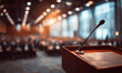 © Shahazadi - A wooden podium with a microphone stands at the front of a large auditorium filled with people in the background, illuminated by warm lighting from overhead.