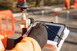 © Iryna - Close-up of construction worker in a bright orange uniform is focused on operating a tablet while standing near traffic cones and surveying equipment on construction site