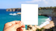 © mienakae - Hand holding a blank instant photo frame with a transparent background for easy overlay. The backdrop is a beautiful, blurred coastal scene with a turquoise sea, evoking travel memories.