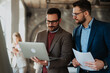 © Novak - Male coworkers talking about business strategy while their team member works behind them in a bright office space