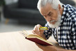 © New Africa - Senior man using magnifying glass while reading book at wooden table indoors, space for text
