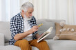 © New Africa - Senior man using magnifying glass while reading book on sofa indoors, space for text