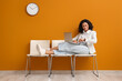 © Pixel-Shot - Female African-American job applicant with wristwatch and laptop waiting for interview near orange wall
