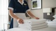 © Eve - Hotel housemaid in navy uniform holding stack of fresh white laundered bedsheets in luxury bedroom. Focus on hospitality service, hygiene standards, and professional housekeeping maintenance.