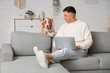 © Pixel-Shot - Young man with modern laptop sitting on sofa and cute Australian Shepherd dog in living room