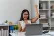 © Kritdanai - Excited Professional Celebrates Success: A young professional celebrates her success, punching the air in jubilation while working at her desk, as a laptop sits open in front of her.
