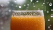 © Thares2020 - Close up of a refreshing orange beverage in a glass covered in condensation viewed through a window on a rainy day