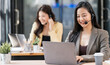 © NAMPIX - Smiling Asian woman with headset working on laptop in modern office, providing customer support with a friendly colleague in the background.