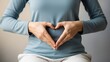 © Amri - Woman wearing blue shirt making heart shape with hands on her stomach against neutral background.