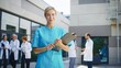 © VAKSMANV - Caucasian female nurse standing outside clinic. Professional holding clipboard confidently. Medical worker focusing on camera. Background doctors engaging in discussion near hospital building.
