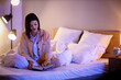 © Pixel-Shot - Beautiful young woman reading book and sitting on bed in bedroom in evening