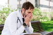 © StockMe - Stressed male doctor sitting outdoors, covering his face with hands, expressing exhaustion and emotional burnout from long shifts and workplace pressure at the hospital