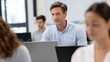 © CYBERPINK - Man in a blue collared shirt sitting in front of a group of people in a meeting room. he is looking at a laptop screen and appears to be engaged in a conversation with the people around him.