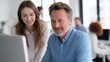 © CYBERPINK - Man and a young woman sitting in front of a computer monitor in an office. the man is on the right side of the image, wearing a blue button-down shirt and has a beard and mustache.