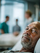 © Daria - Elderly man with gentle expression resting in hospital bed with blurred medical staff attending to background in modern healthcare facility environment
