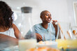 © Nexa - Smiling Black woman with shaved head eating a snack and laughing with a friend in a bright kitchen.