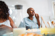 © Nexa - Cheerful Black woman with a shaved head laughing and eating breadsticks with a friend during a meal at home.