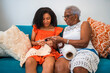 © Nexa - African American grandmother teaching her teenage granddaughter how to crochet while sitting together on a sofa at home.
