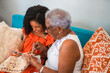 © Nexa - Happy African American grandmother and granddaughter laughing while crocheting together on a blue sofa, sharing a joyful moment of intergenerational bonding at home.