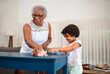 © Nexa - Happy African American grandmother and granddaughter cleaning a blue table together at home, sharing a moment of bonding and teaching household chores.