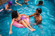 © Nexa - Happy multi-ethnic friends enjoying a summer pool party with colorful inflatables, focusing on a young couple laughing together in the blue water.