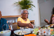 © Nexa - Smiling elderly Black woman sitting at a dining table with family, reaching for food during a meal at home.