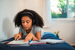 © Nexa - Focused teenage girl with curly hair wearing headphones while studying and writing in notebooks on her bed at home.