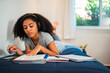 © Nexa - Focused teenage girl with curly hair wearing headphones and studying with books while lying on her bed at home.