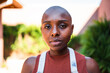 © Nexa - Portrait of a confident young Black woman with a shaved head looking directly at the camera in an outdoor setting.