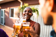 © Nexa - A cheerful Black woman with a shaved head celebrates with friends, clinking beer glasses during an outdoor social gathering at sunset.