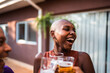 © Nexa - Happy young Black woman with shaved head laughing and clinking beer glasses with friends at an outdoor backyard party.