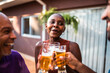 © Nexa - Joyful black woman with shaved head celebrating and clinking beer glasses with friends during a summer backyard gathering.