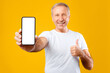© Prostock-studio - A man stands in front of a yellow background holding a smartphone with a blank screen. He smiles widely and gives a thumbs up, conveying a sense of excitement and approval.