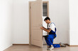 © Prostock-studio - A young locksmith in a blue uniform is kneeling as he installs a doorknob handle on an entrance door. This professional repair service ensures the door is secure and functional.
