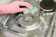 © Amore Sea - Hand in pink glove scrubbing gas burner on stainless stove with soapy sponge. Kitchen appliance cleaning maintenance.